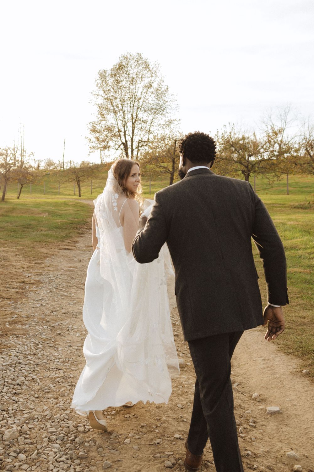 Bride in flowing white dress and groom in dark suit walk together on dirt path through autumn countryside near vernon nj.