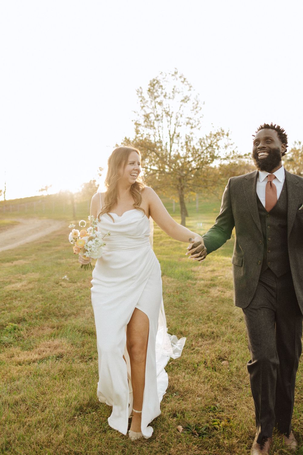 A joyful wedding couple holding hands and walking through a sunlit field at golden hour in a farm near vernon nj.