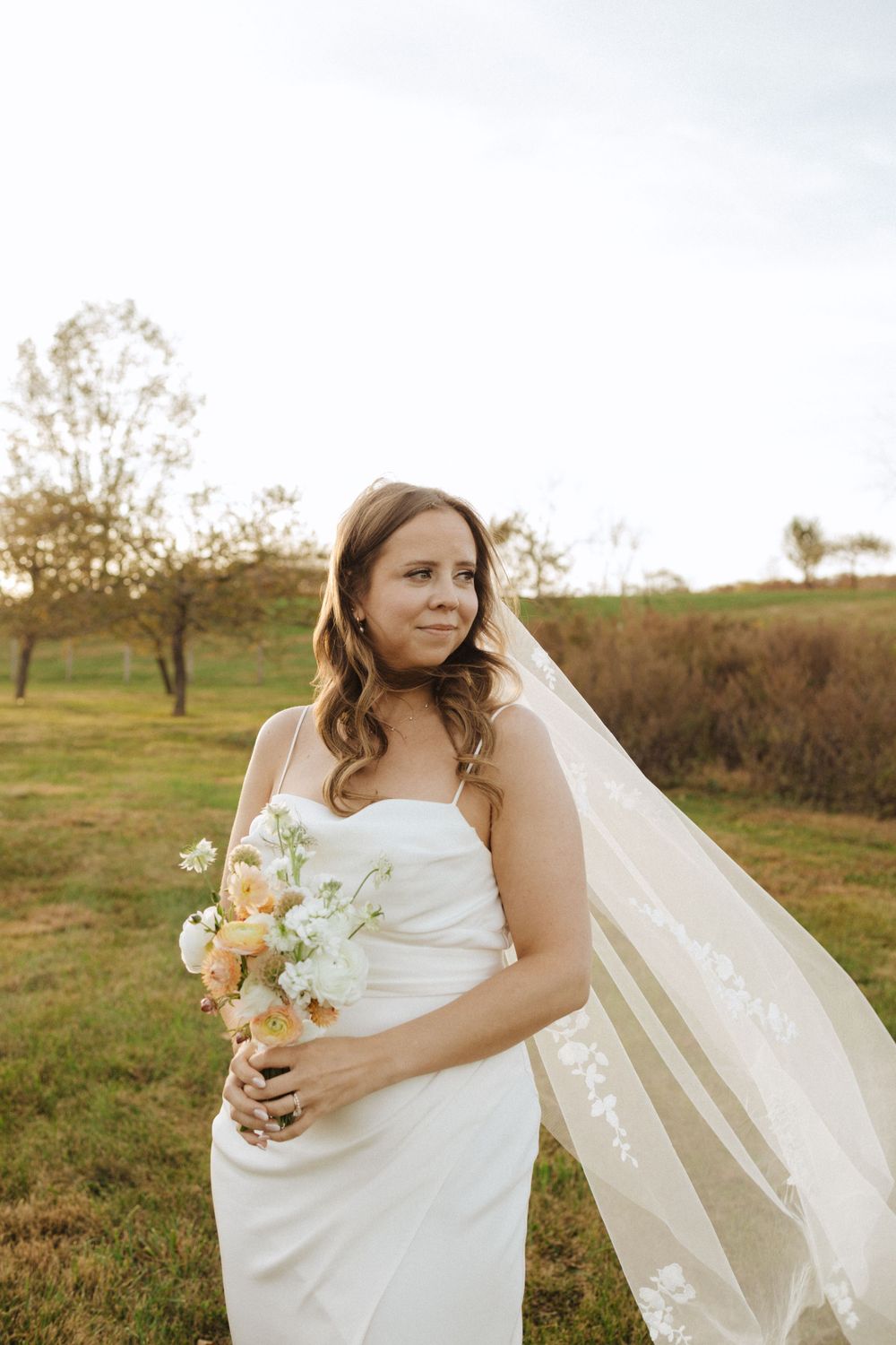 A bride in a white satin gown holds a romantic pastel bouquet in a rustic outdoor setting near vernon nj.