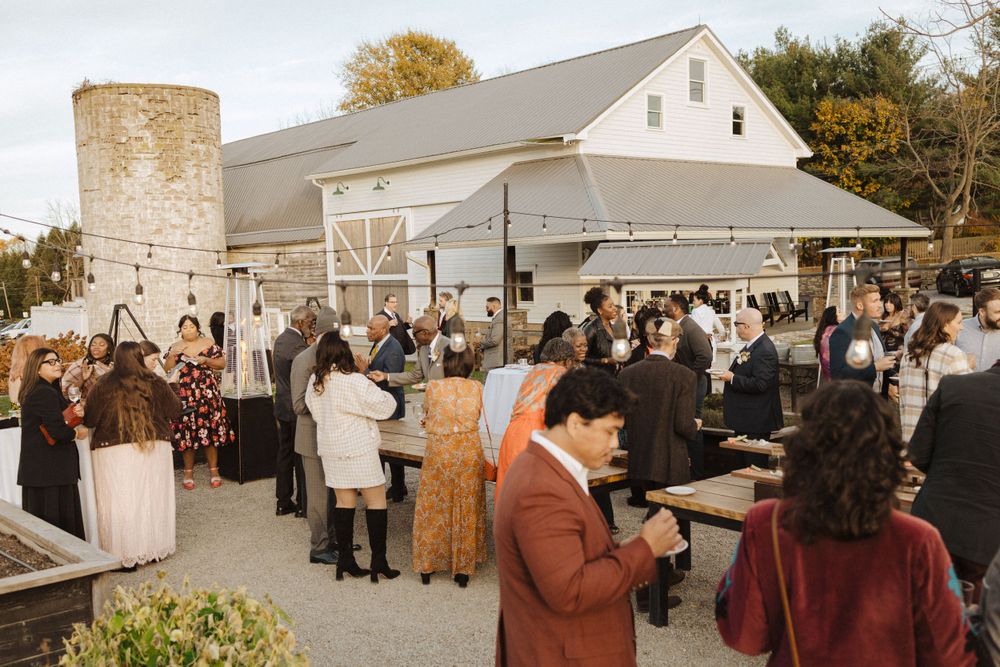 Guests mingle at an outdoor wedding reception near a rustic white barn with stone chimney during golden hour near vernon nj.