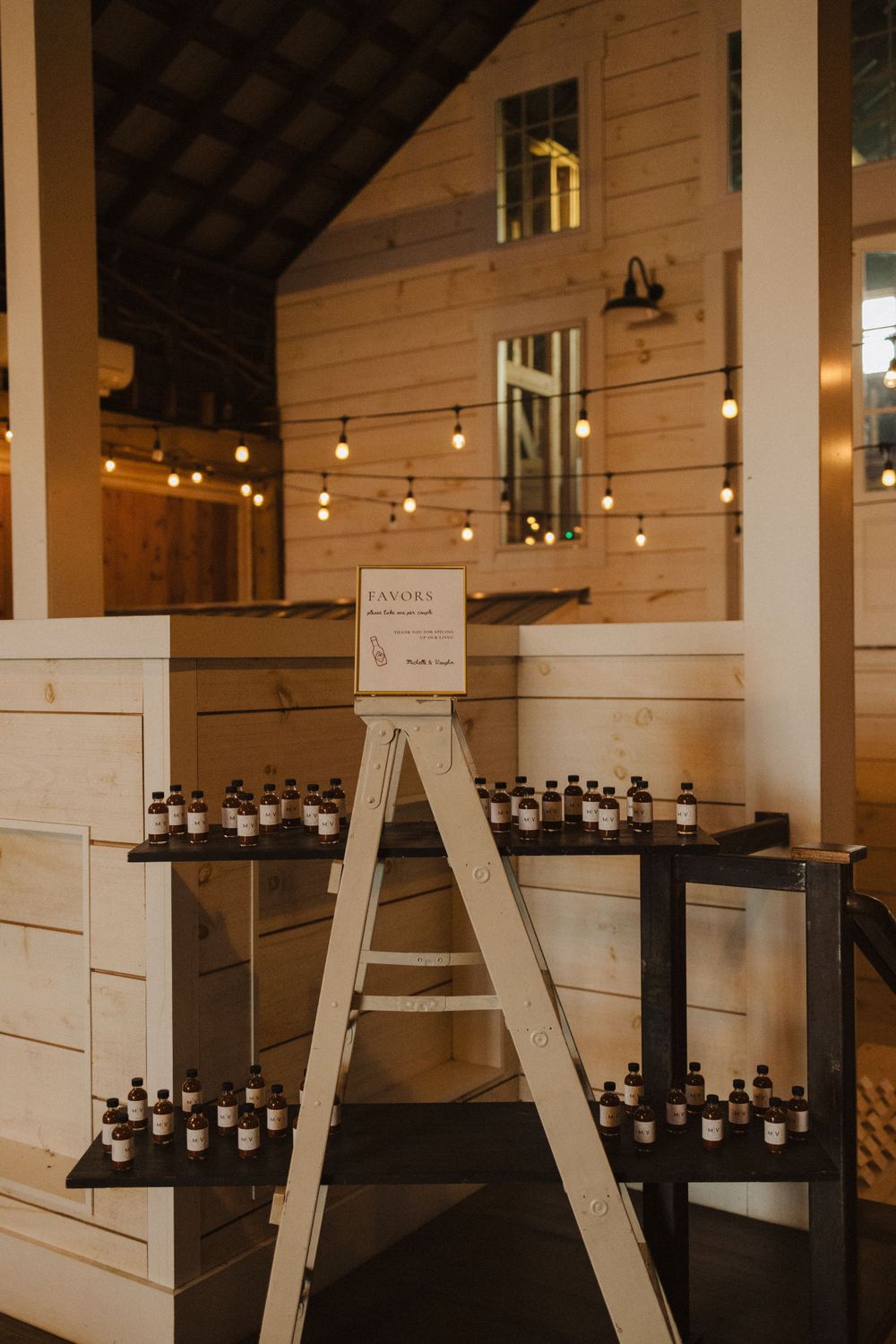 A rustic white wooden ladder display with rows of small dark bottles and string lights in a barn-style venue near vernon nj.