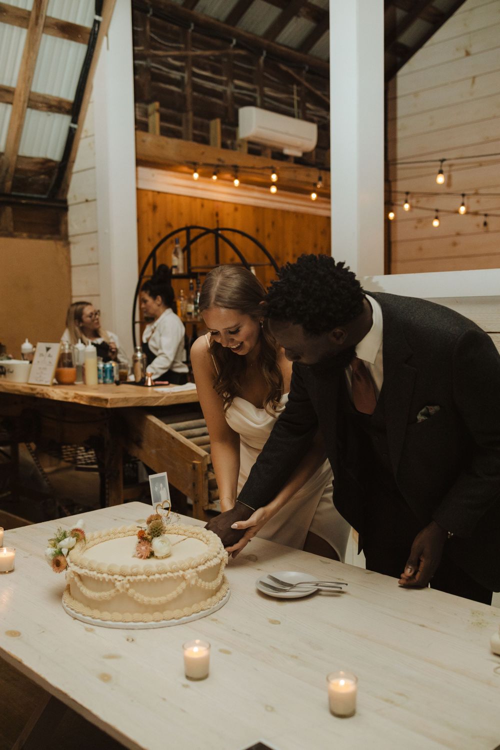 A couple cuts into their classic white wedding cake at a rustic barn reception decorated with string lights near vernon nj.