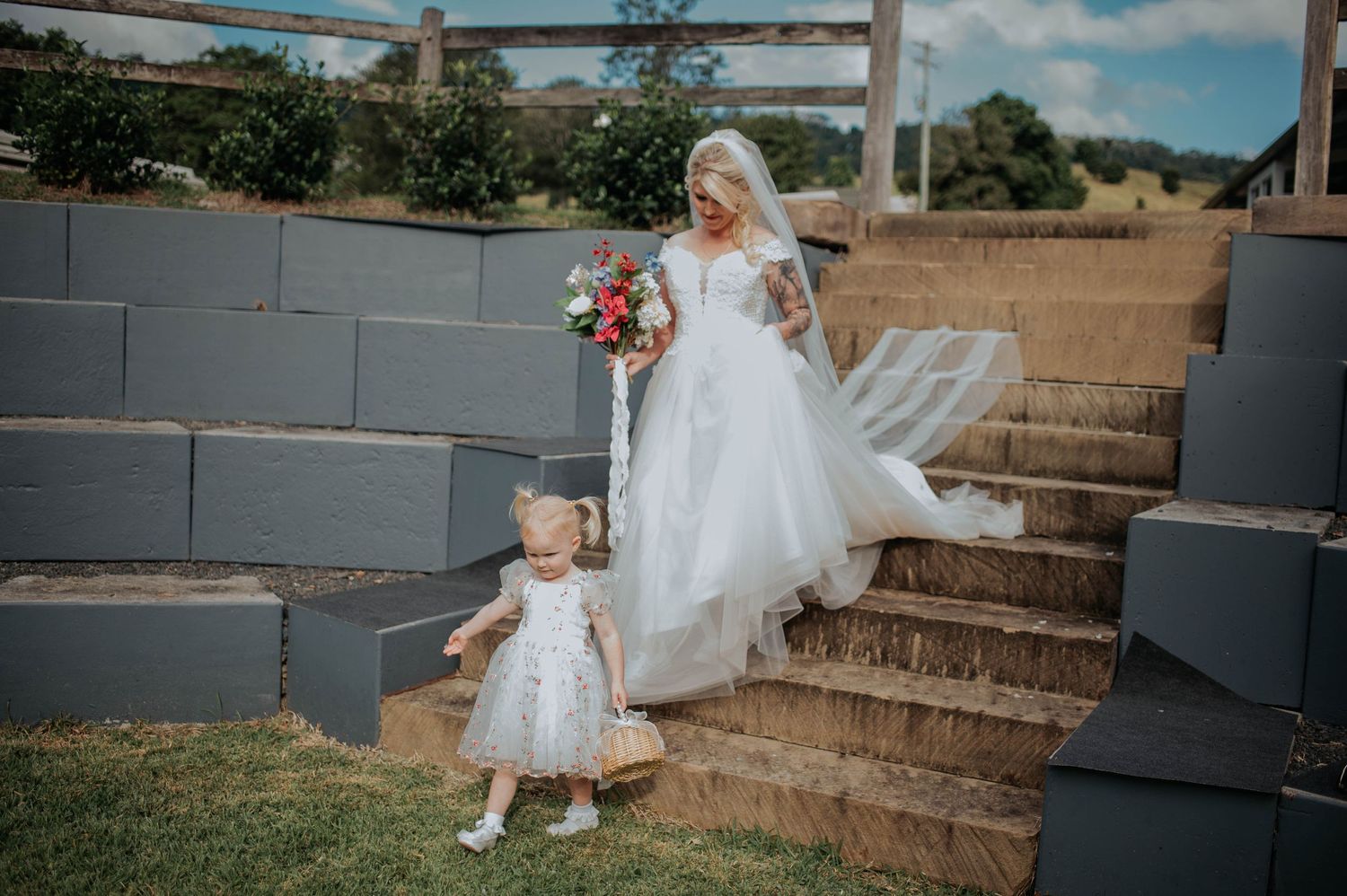 A bride in white gown descends stone steps with a small child in a matching dress.