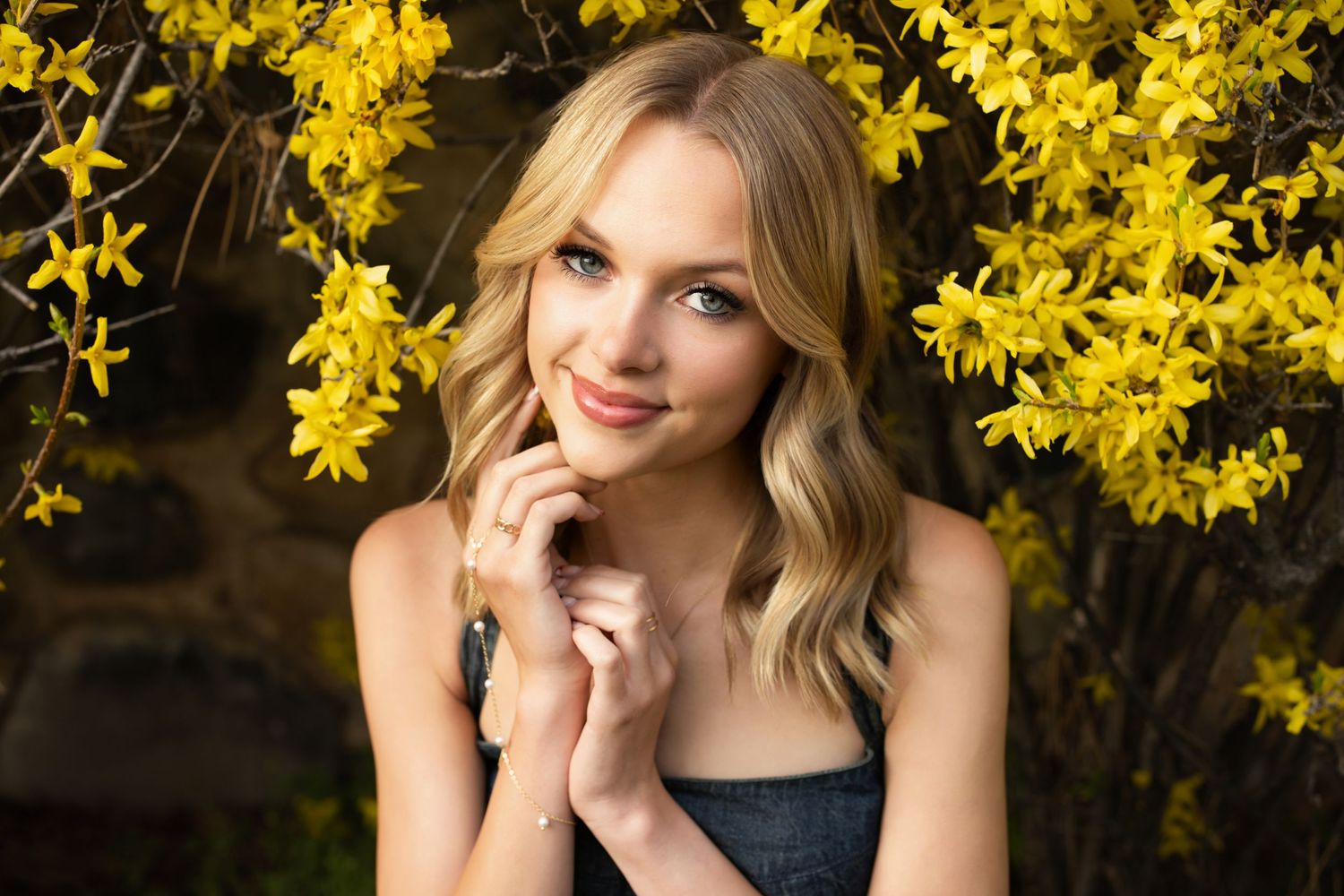 A blonde woman in a black top poses outdoors against a backdrop of bright yellow spring flowers.