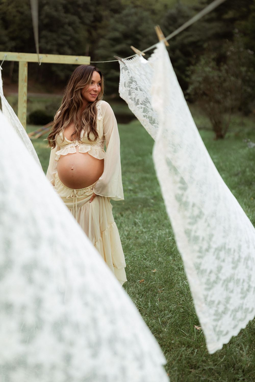 Pregnant woman in sheer yellow dress standing outdoors among hanging white lace fabric on a clothesline.