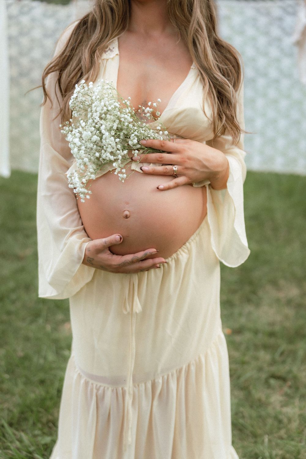 Pregnant woman in cream dress holding baby's breath flowers against her bare belly outdoors.