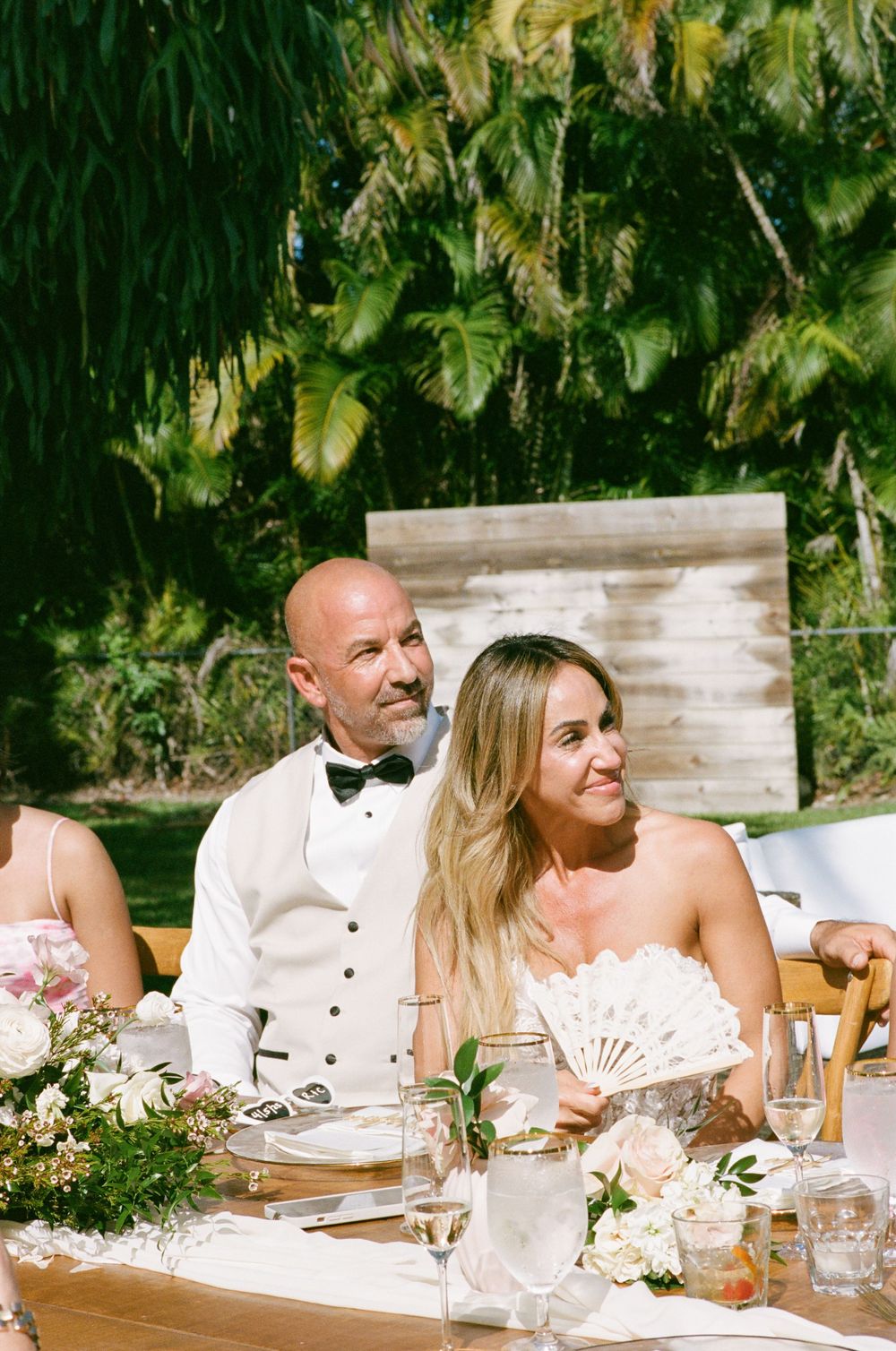 A couple sit at an elegantly decorated outdoor wedding reception table with floral centerpieces and glassware.