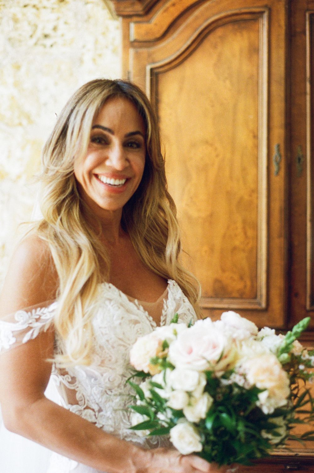 A bride in a lace wedding dress holds a romantic bouquet of white and pink roses against a wooden door backdrop.