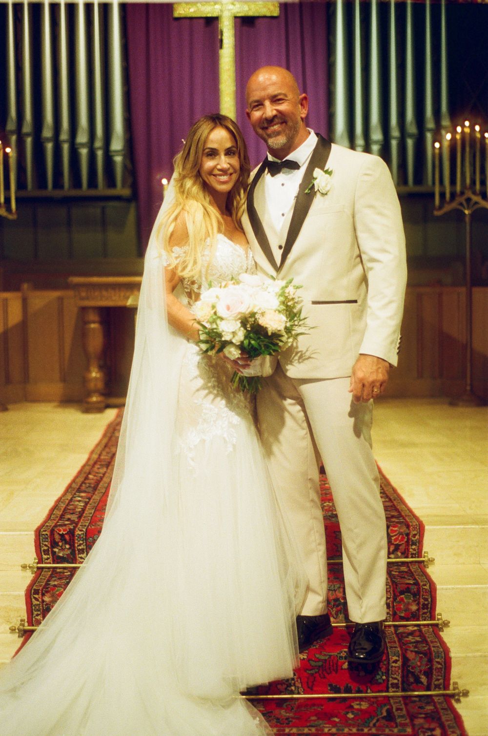 A bride and groom stand together at a church altar with organ pipes and a cross visible in the background.