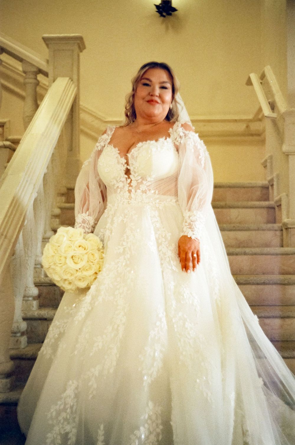 A bride wearing an elegant white lace wedding gown with long sleeves holds a white rose bouquet on a grand staircase.
