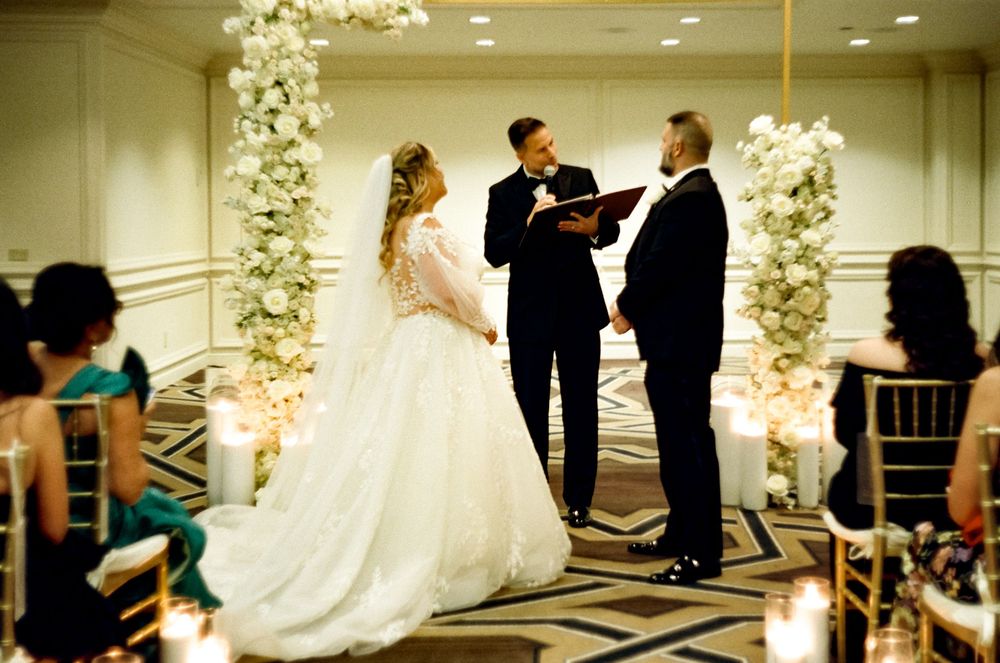 Wedding ceremony takes place under beautiful floral arch with white orchids in an elegant indoor venue.