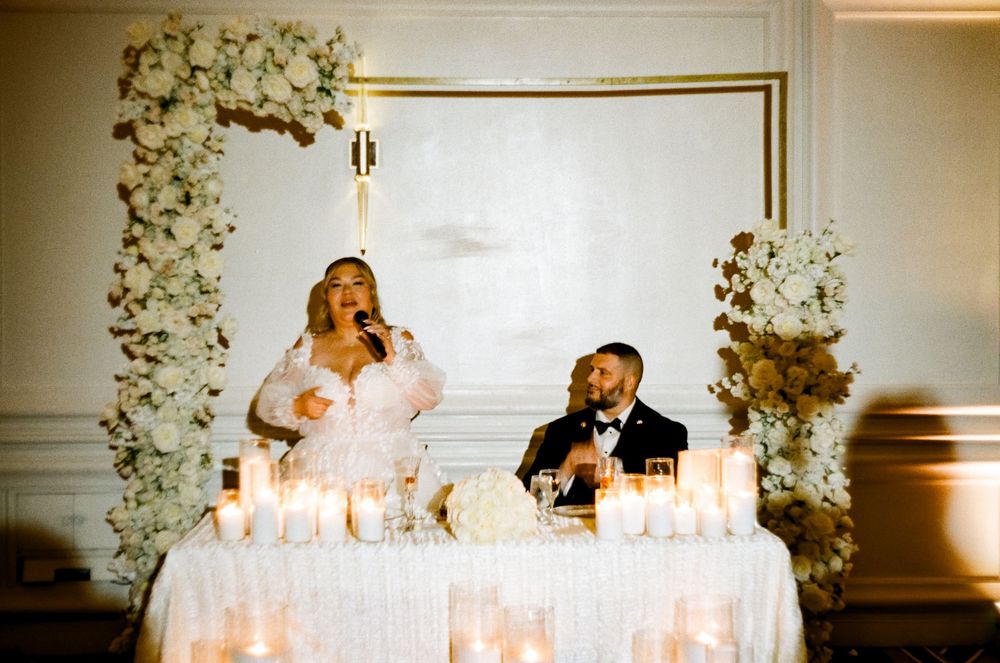 A couple sits at an elegant wedding reception table with floral arch decorations and glowing candles at a formal venue.