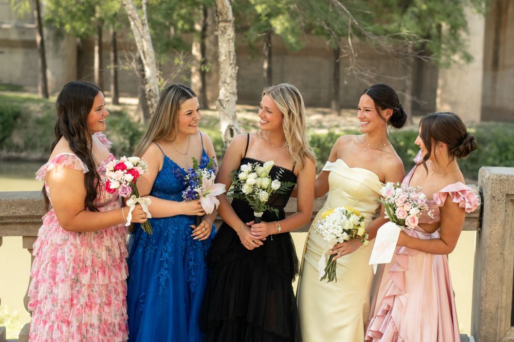 A group in formal attire stands together outdoors wearing colorful dresses in pink, blue and black while holding bouquets.