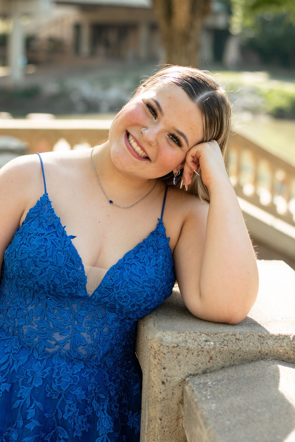 Smiling person in a royal blue lace formal dress leaning on stone railing in outdoor sunlit setting.