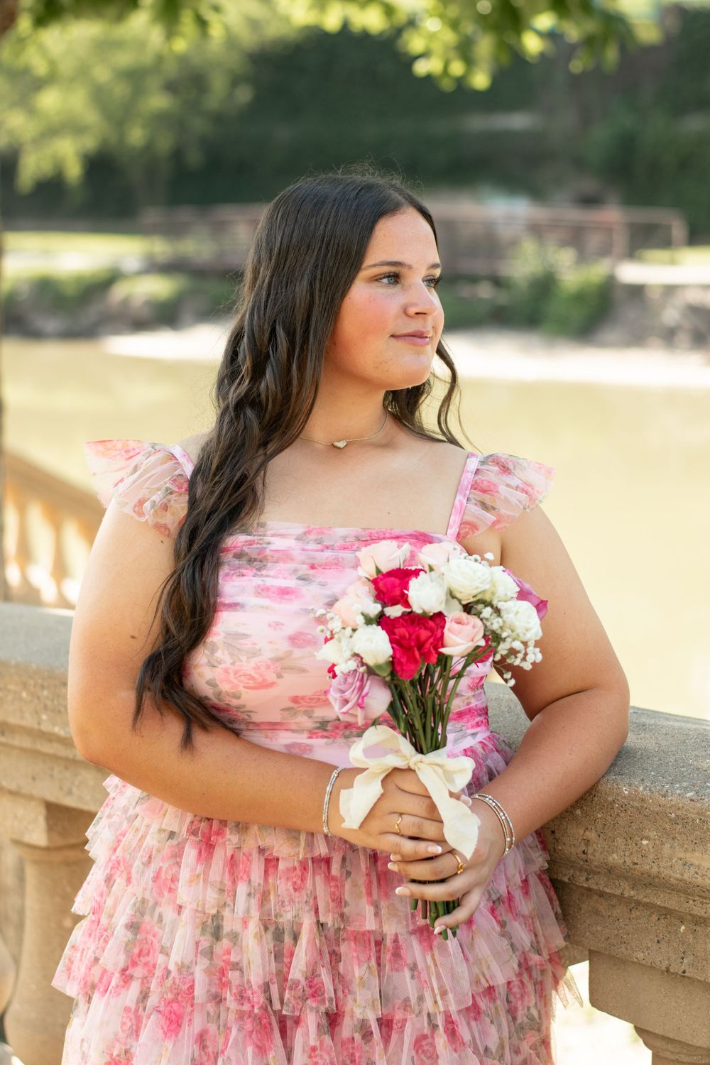 A person in a pink floral dress holding a bouquet of red and white flowers poses by a stone railing in a garden setting.