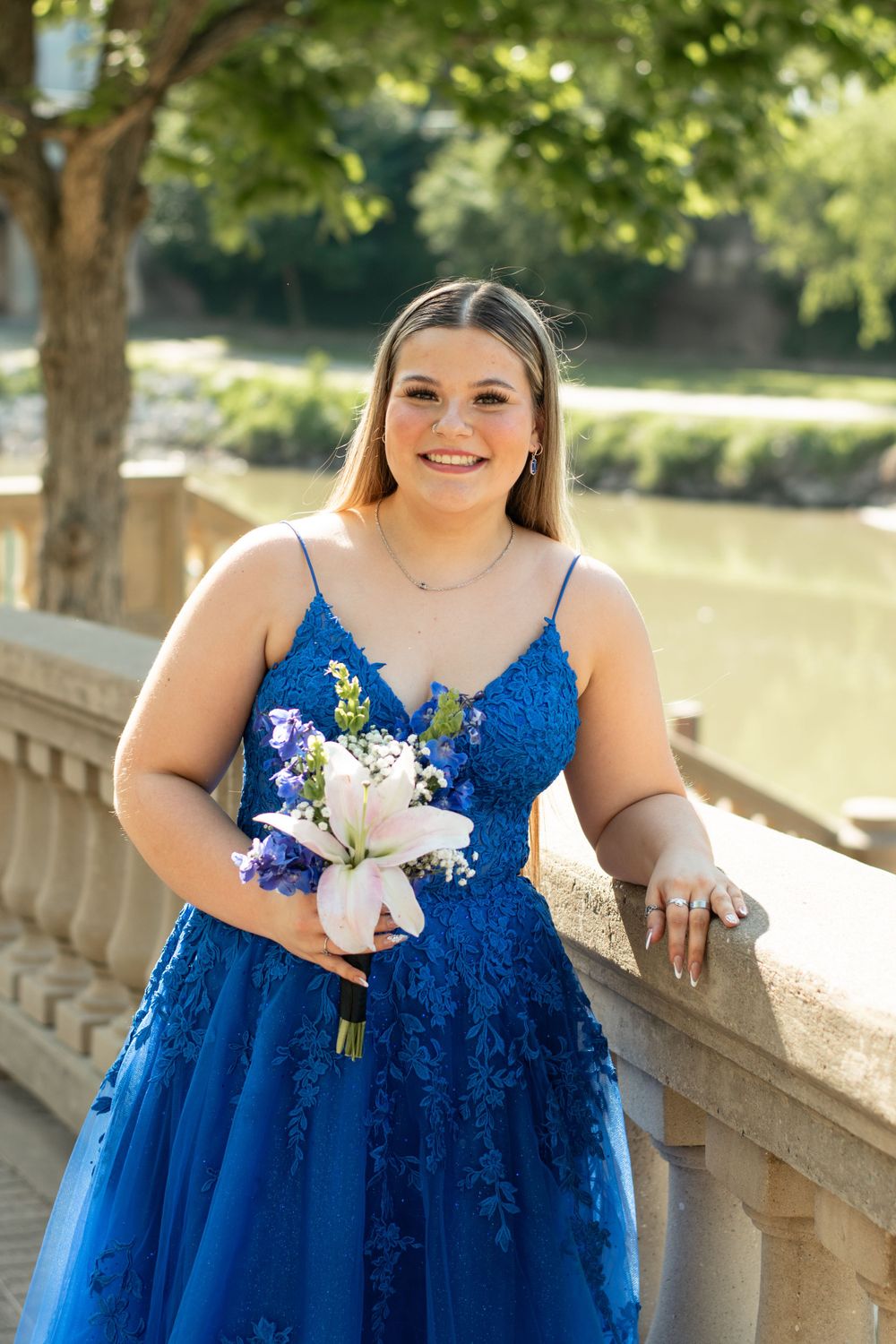 Beautiful formal dress in royal blue lace and tulle paired with a white and purple floral bouquet by stone railing outdoors.