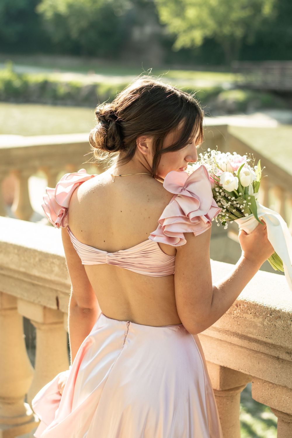 A person in a pink ruffled dress with a low back holds a bouquet while standing at a stone balustrade in sunlit garden.