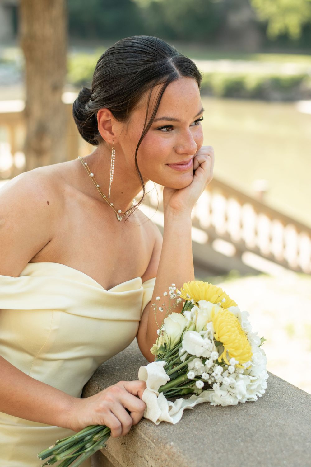 A person in a cream strapless dress holds a bouquet of yellow and white flowers while posing outdoors.