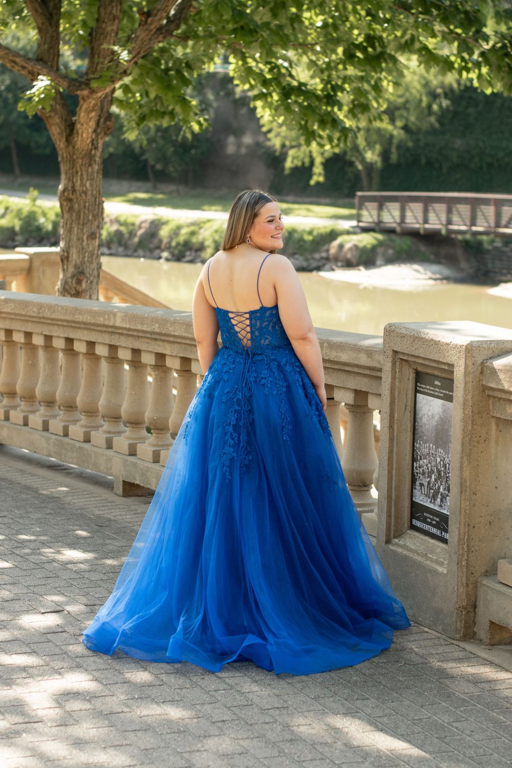 A person wearing a royal blue ball gown with corset back stands on a stone bridge in a park setting.