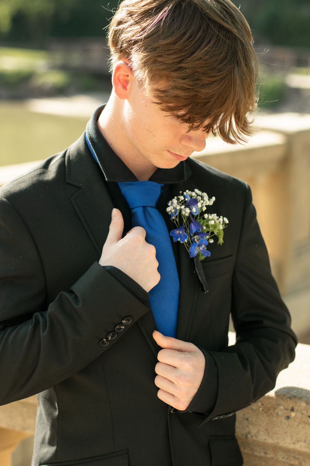Person wearing black suit with bright blue tie and delicate blue floral boutonniere adjusts tie in sunlit outdoor setting.