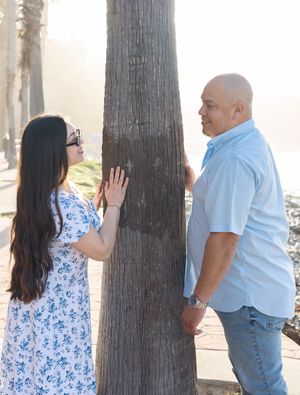 Two people stand on opposite sides of a palm tree trunk on a sunny coastal path.