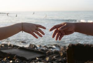 Two hands reach toward each other against a scenic ocean background with rocks and waves at sunset.
