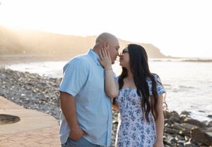 A couple shares an intimate moment on a rocky beach at sunset with mountains in the background.