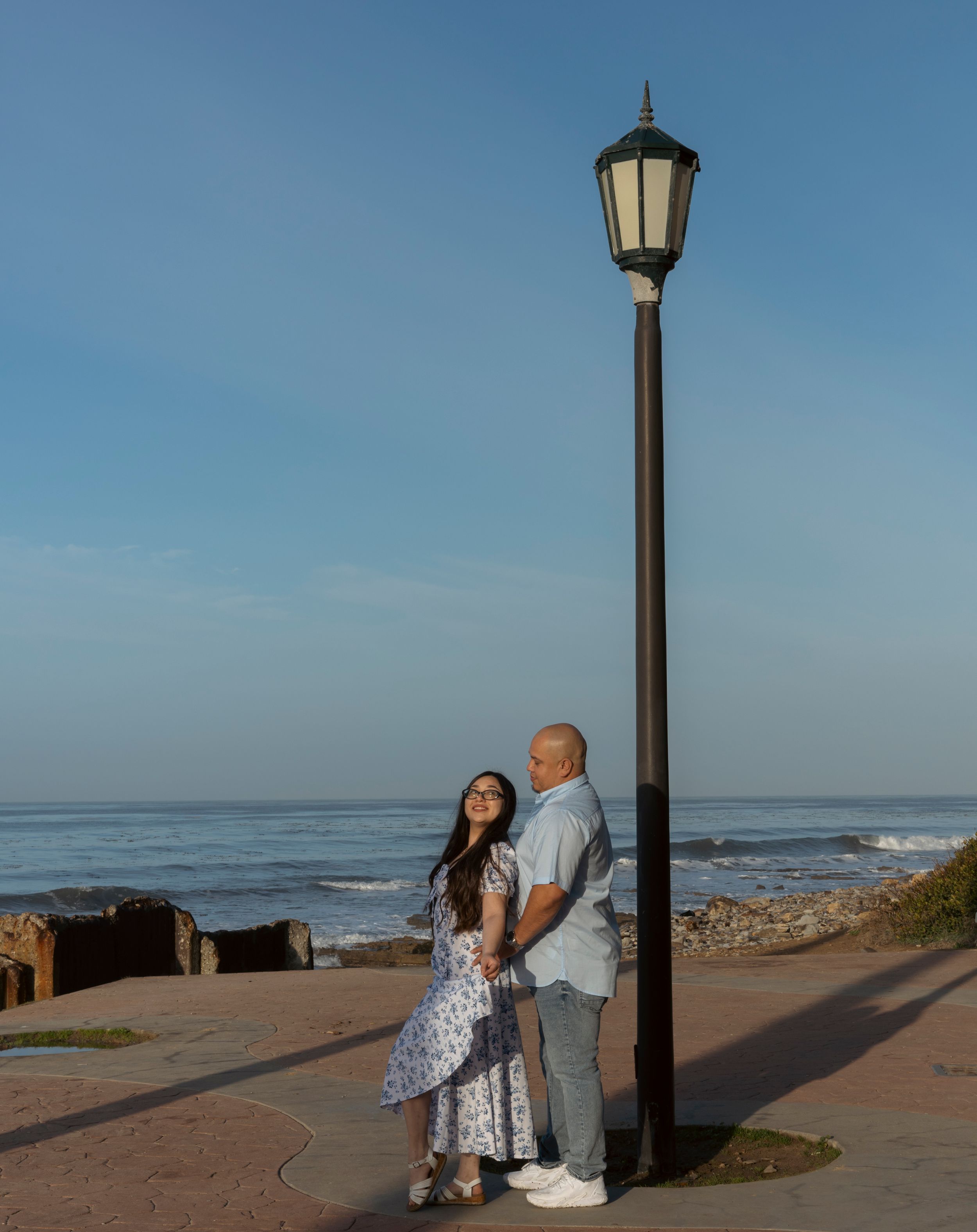 A couple stands together by a vintage lamp post along a coastal pathway during sunset.