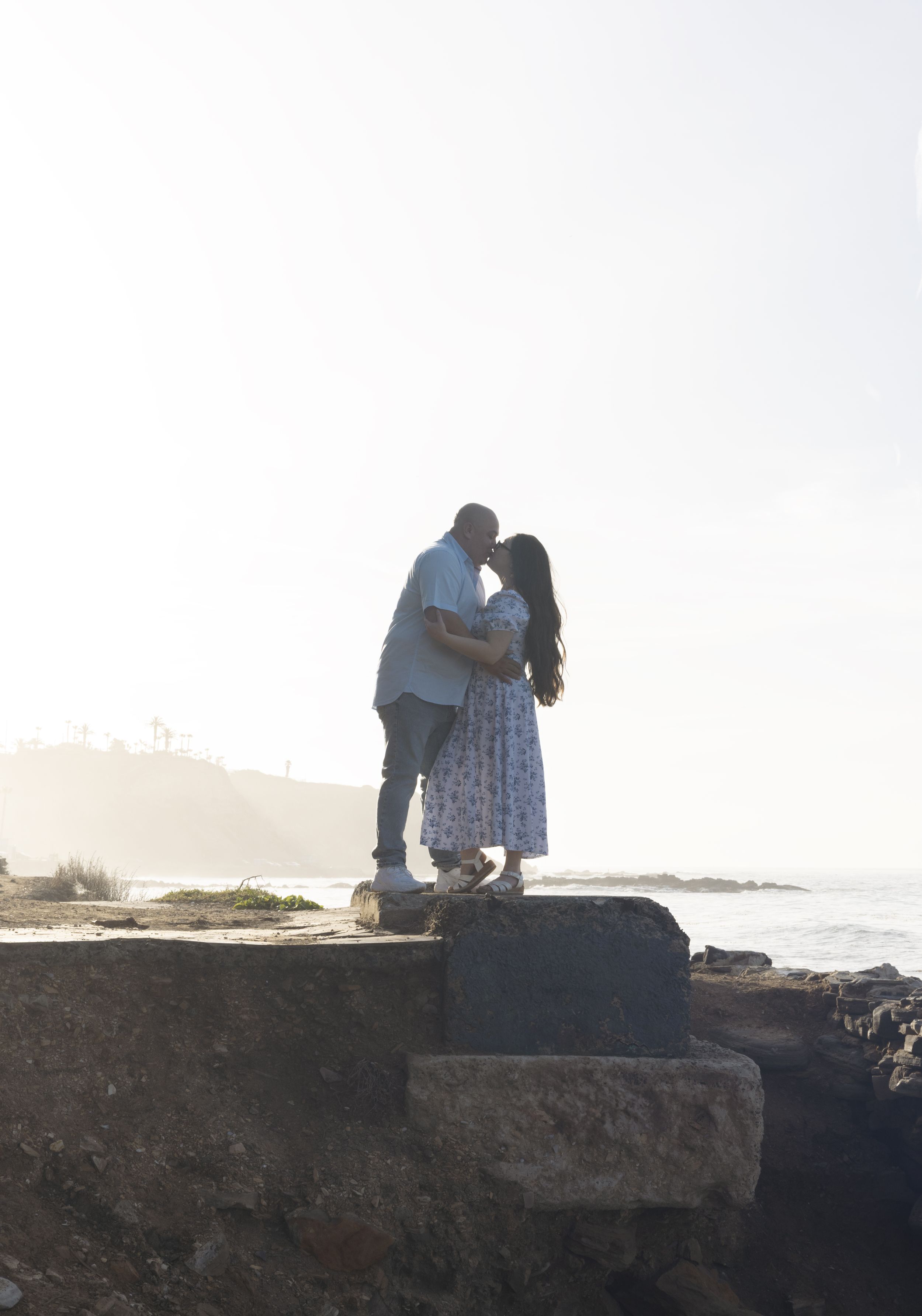 A couple embraces while standing on rocky coastal cliffs at sunset with ocean and misty mountains in the background.