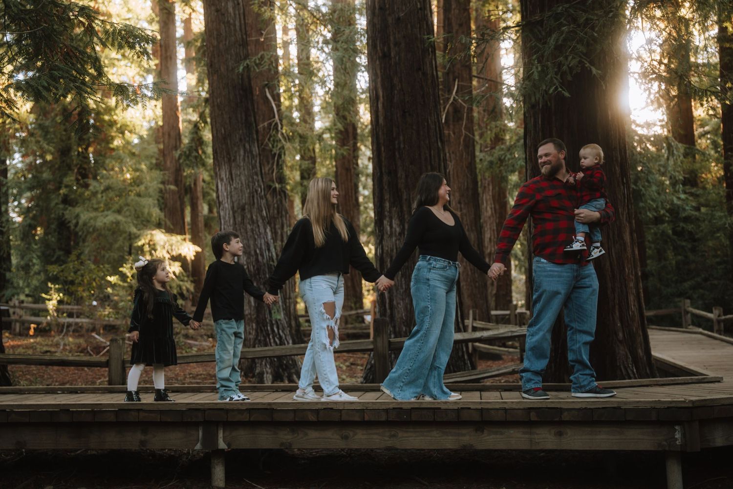 A group of people standing on a wooden walkway surrounded by tall trees in a forest at sunset.