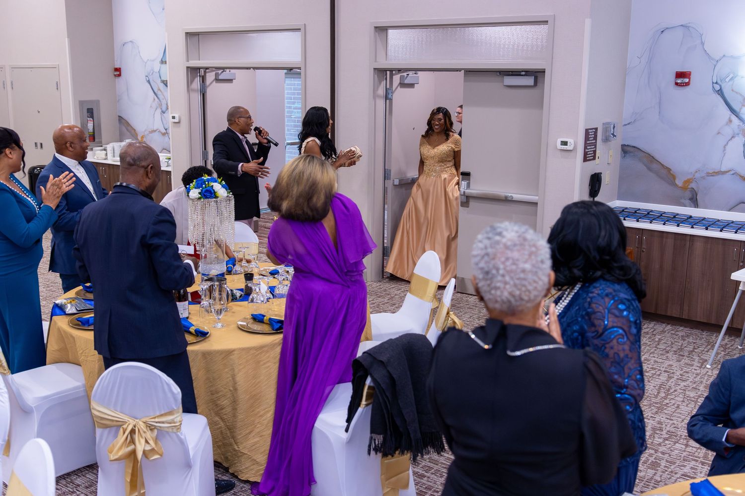 Event guests in elegant formal attire gathered around decorated tables with gold accents at an indoor banquet venue.