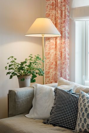 Cozy living room corner with floor lamp, houseplant and decorative throw pillows on gray couch by window with coral curtains.