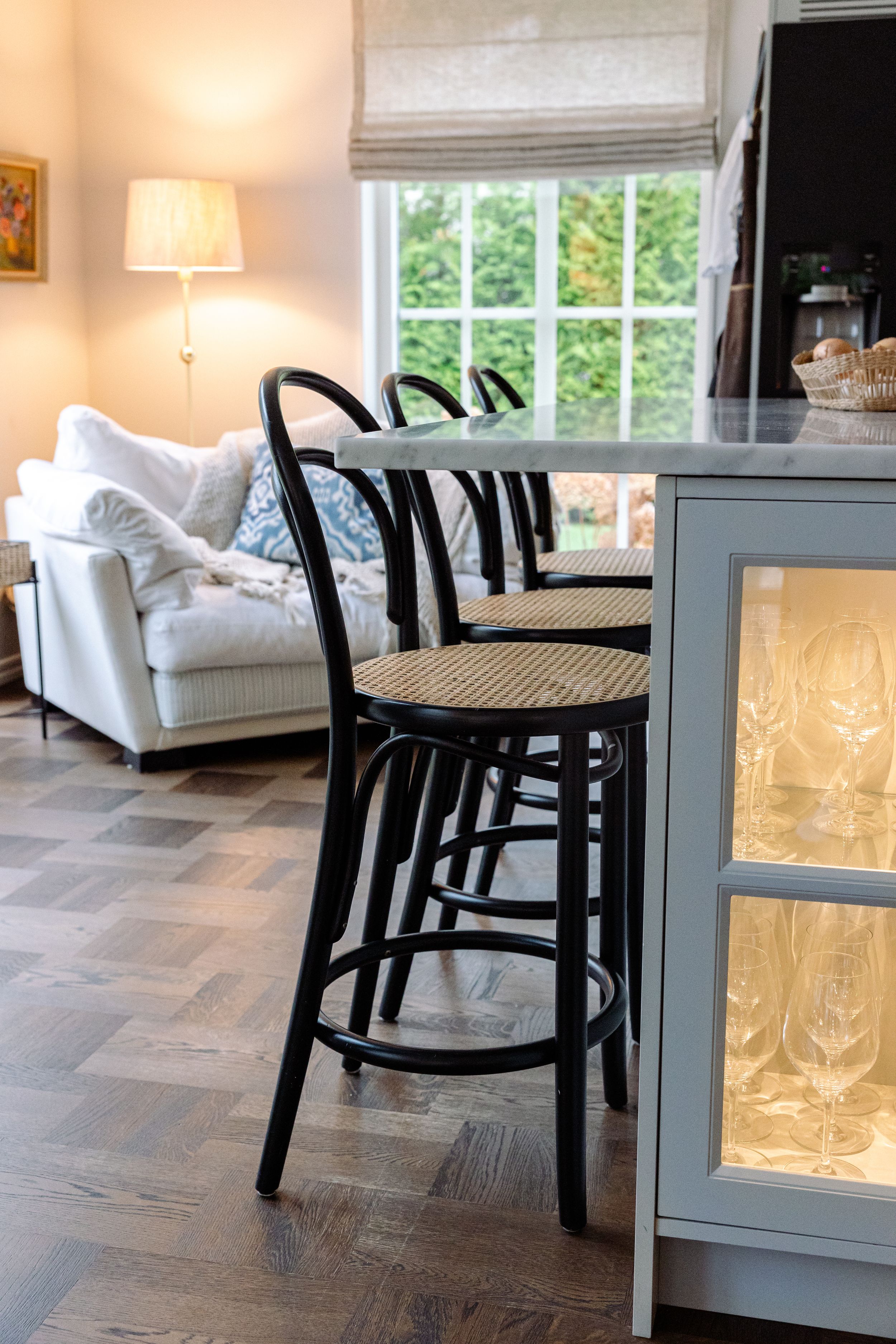 Black wooden bar stools with cane seats line up against a gray cabinet in a cozy room with natural light from windows.