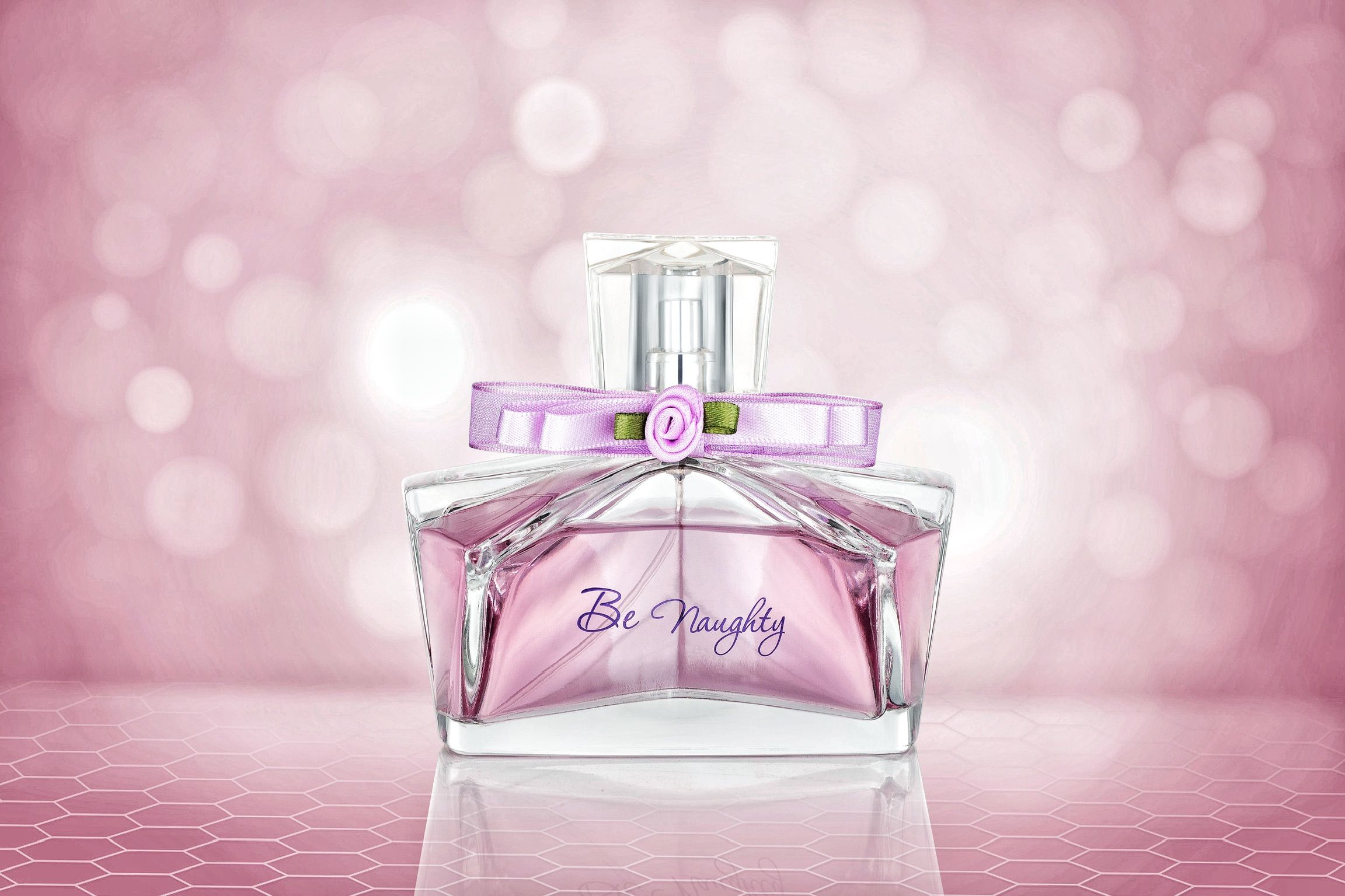 Pink glass perfume bottle labeled Be Naughty with bow detail on sparkling pink bokeh background.