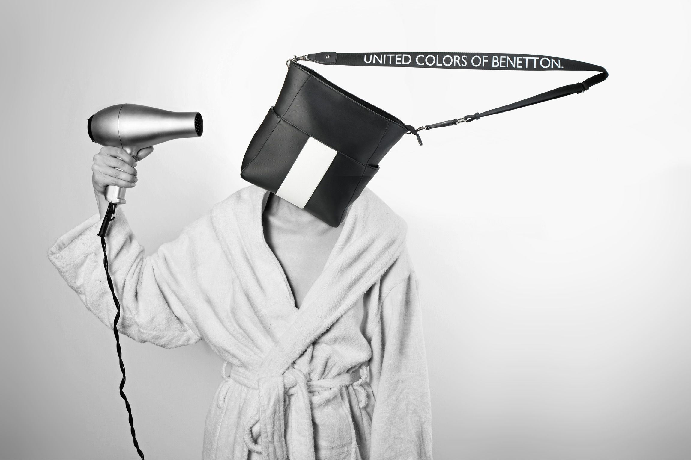 Person in white robe holding hair dryer with United Colors of Benetton bag over their head, black and white photo.