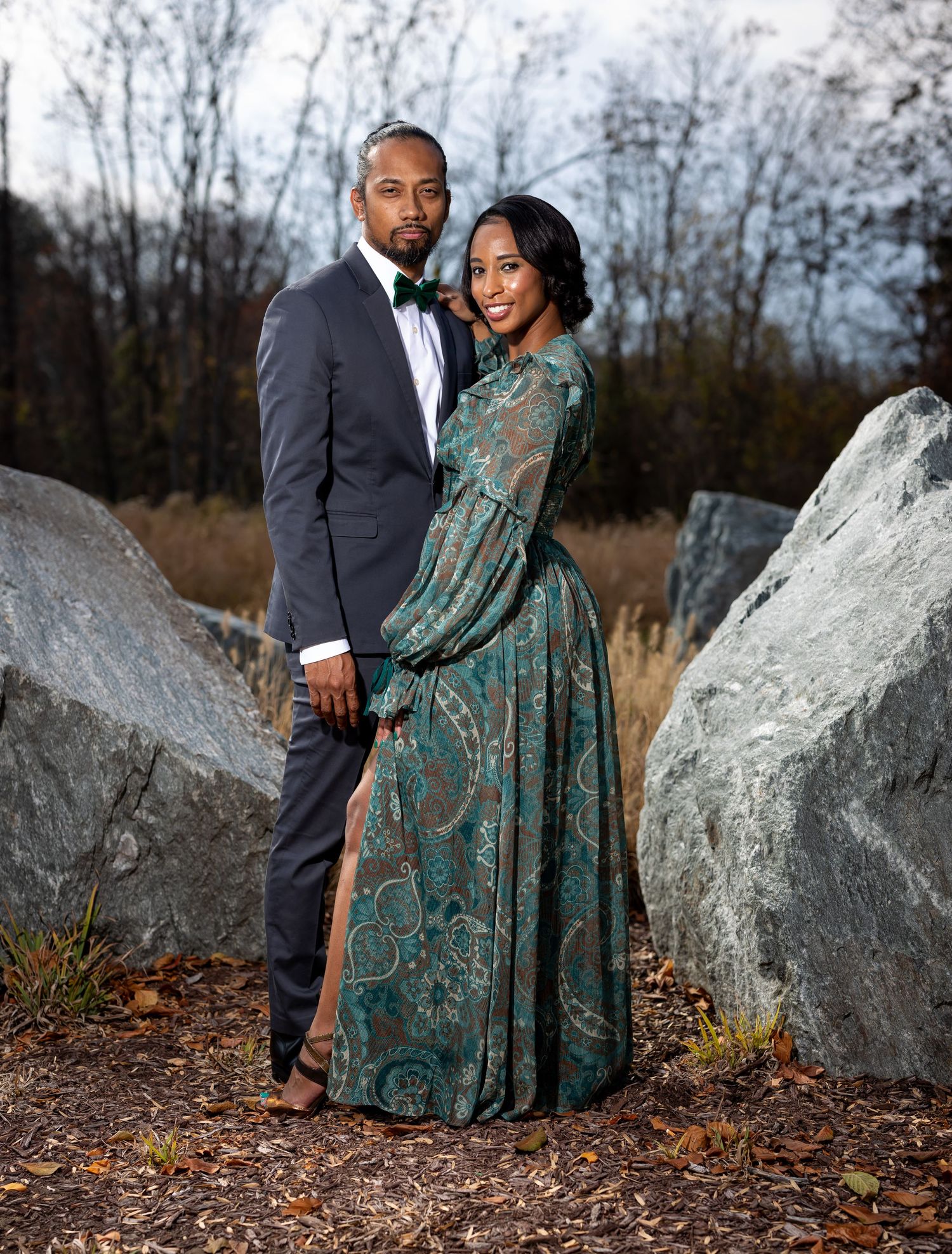 A couple poses outdoors among large rocks, with the woman wearing an elegant teal patterned dress.
