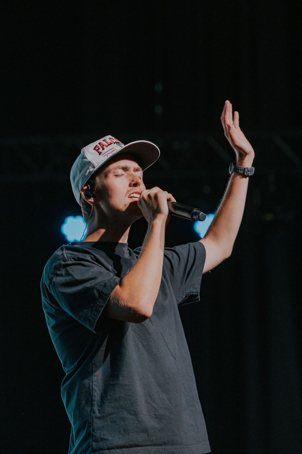 A performer in a baseball cap and dark shirt sings into a microphone during a concert performance.