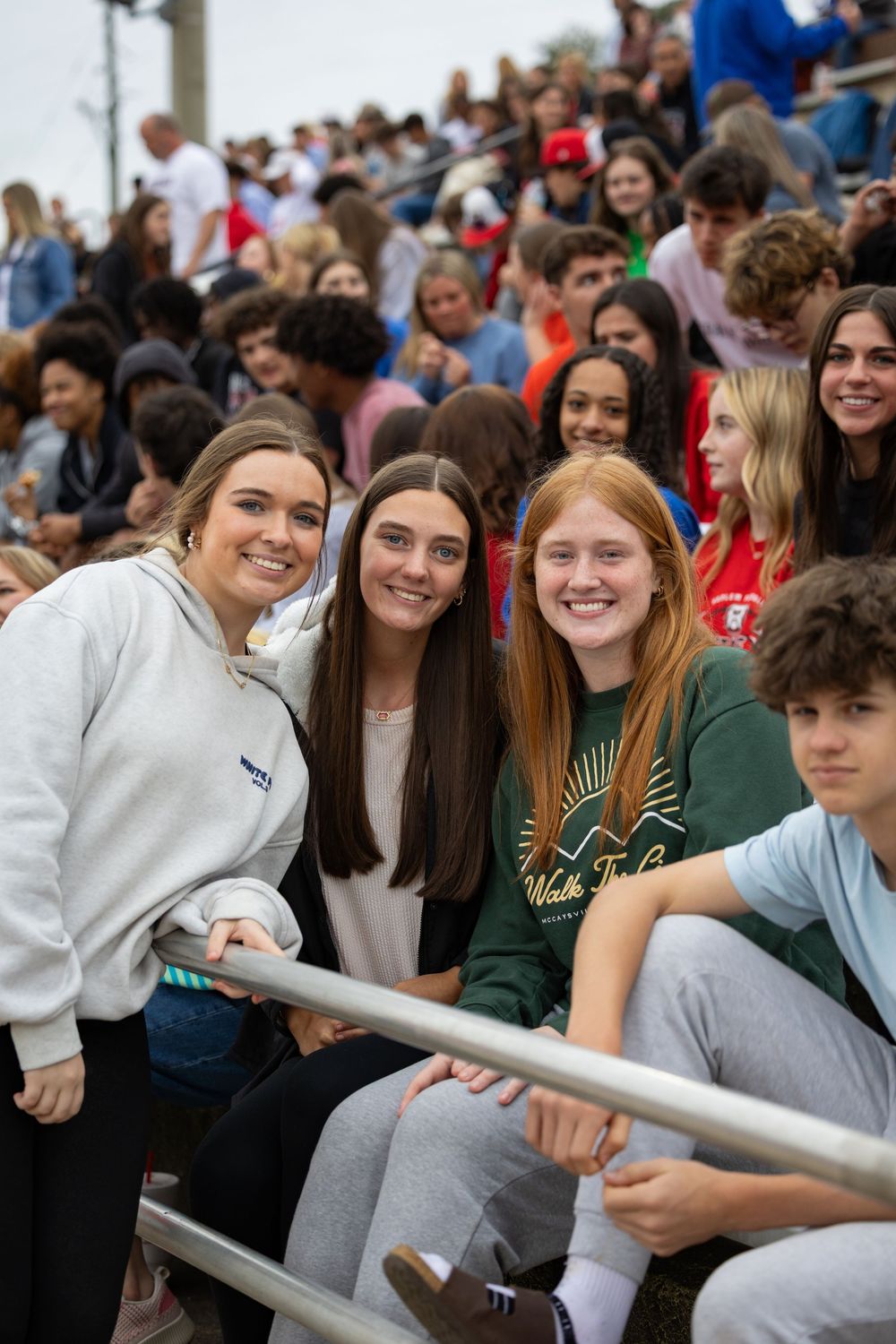 Students sitting together at a crowded school event lean on handrails and smile while enjoying the atmosphere.