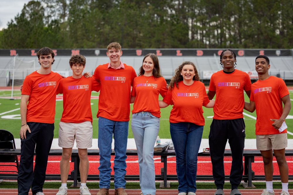Students in matching red t-shirts stand together on a sports field with trees in the background.