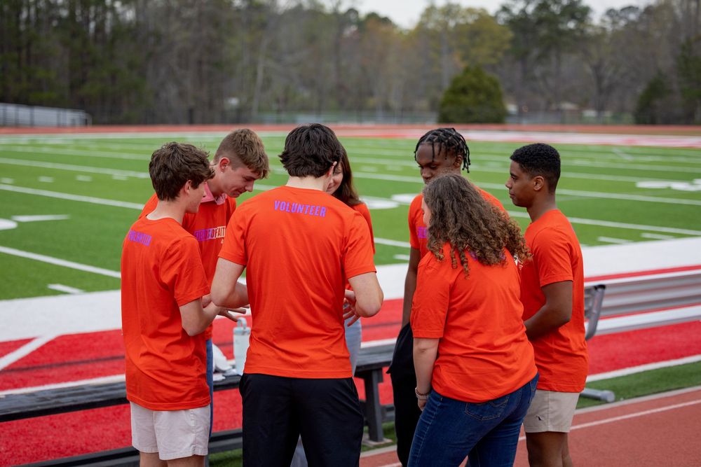 A group wearing orange shirts huddles together on an athletic field with stadium seating in the background.