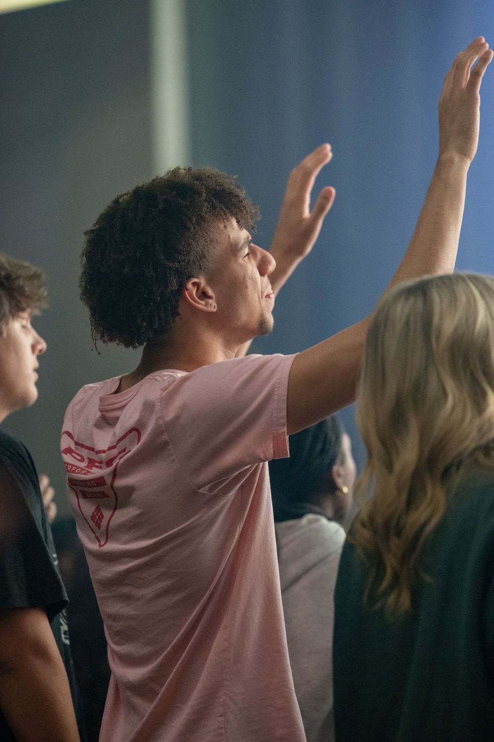 A person in a pink shirt worshipping with raised hands during a group gathering.