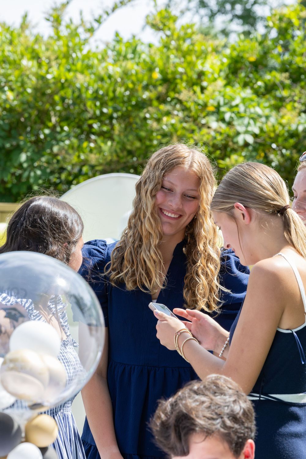 Group of young people gathered outside on a sunny day looking at something on a phone or device together.
