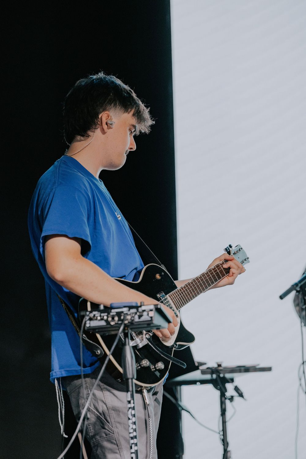 Musician in blue t-shirt playing electric guitar on stage with drum set visible in background.