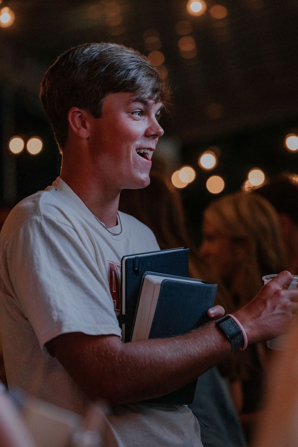 Student holding books smiles while engaging in discussion with blurred lights in background.