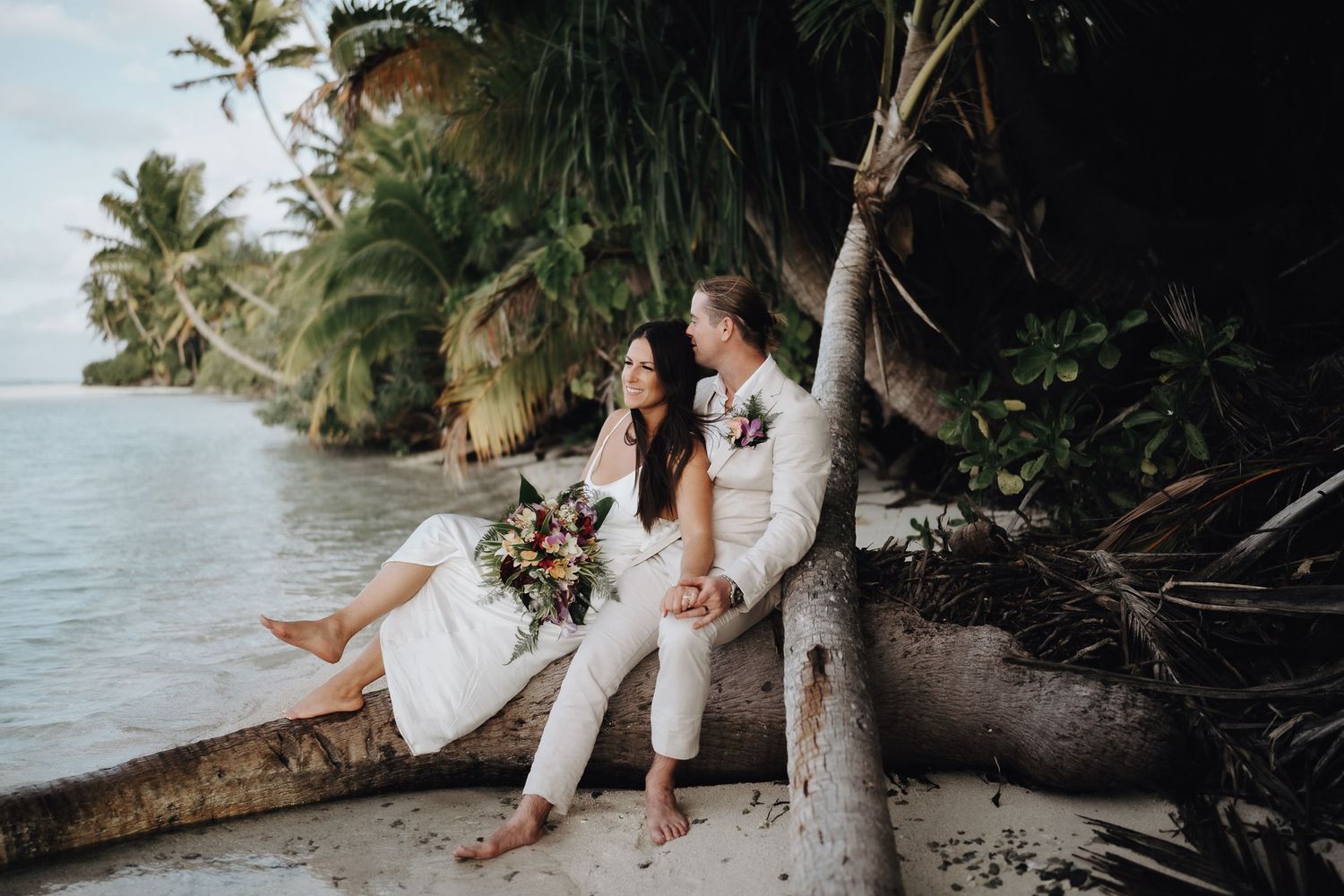 A couple in white outfits sits on a fallen palm tree trunk by the ocean at a tropical beach.