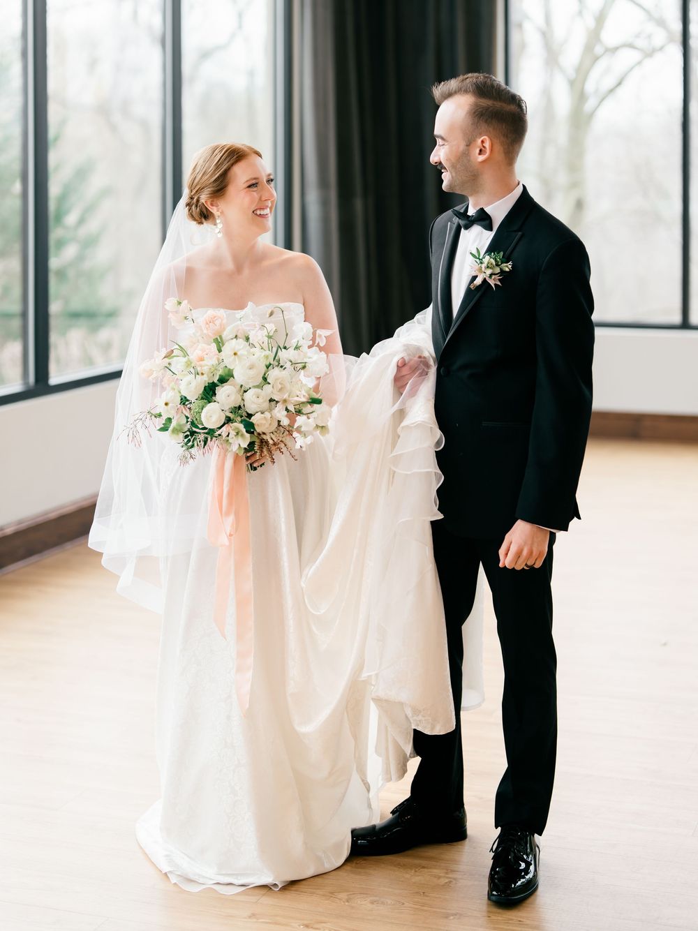 Elegant wedding couple poses together in formal attire near bright windows in a classic indoor venue.