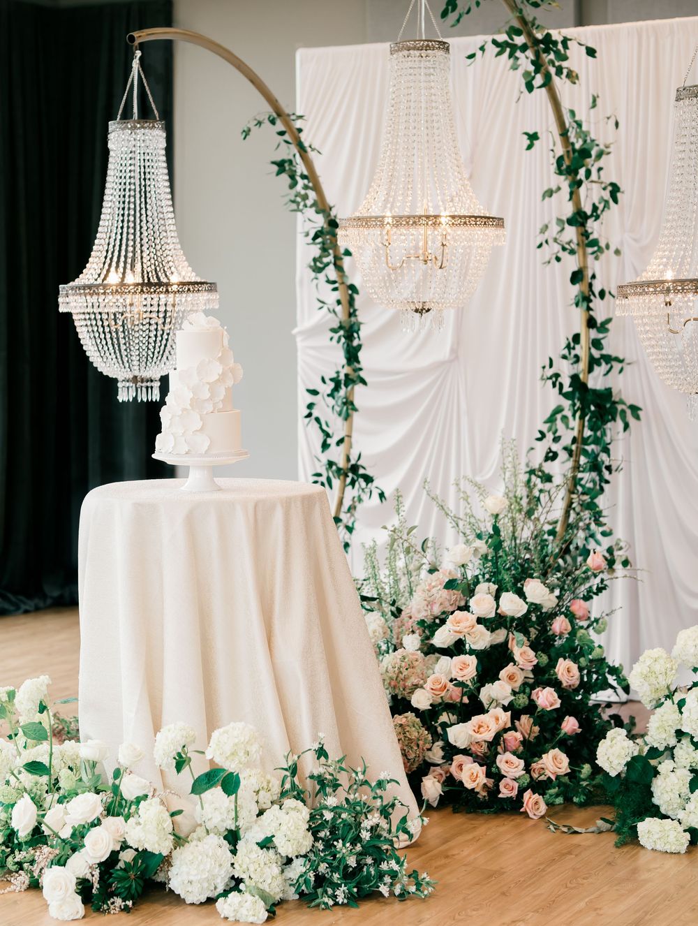 Elegant wedding reception decor features hanging crystal chandeliers, white drapes, climbing greenery, and pink roses.