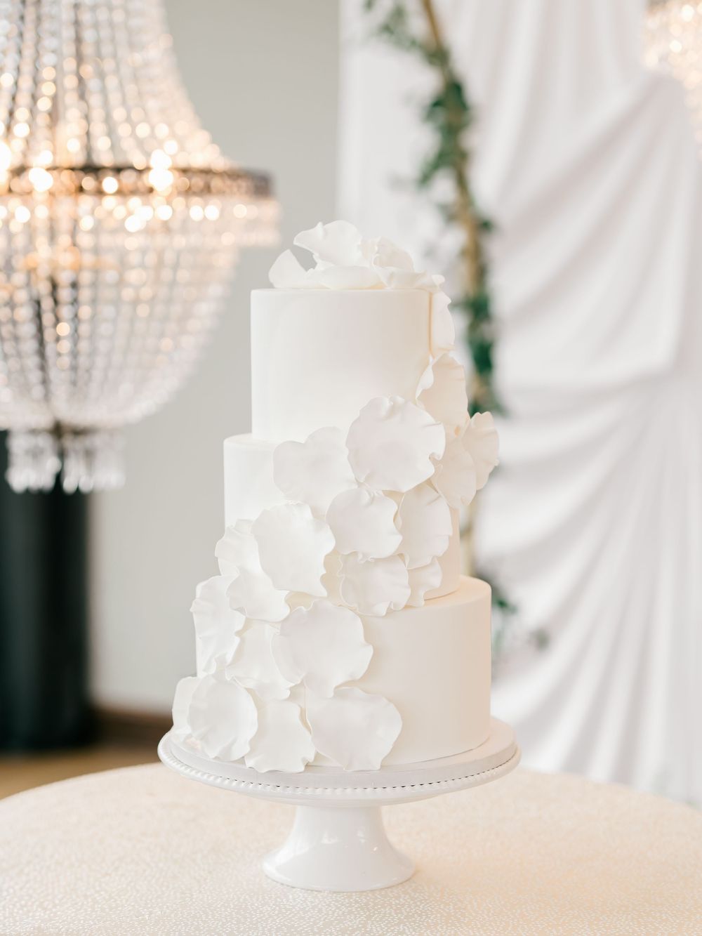 Elegant three-tier white wedding cake with cascading floral details displayed under a crystal chandelier.