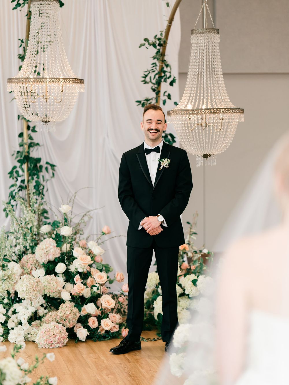 Emotional first look wedding reactions in an elegant ballroom with crystal chandeliers and white floral decorations.