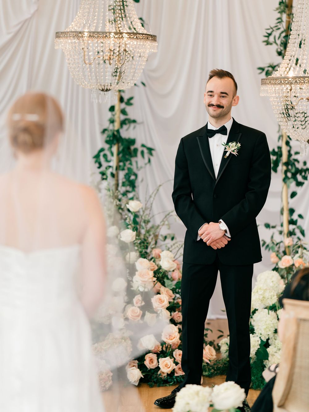 A series of wedding photos showing a first look moment in an elegant venue decorated with chandeliers and floral arrangements.
